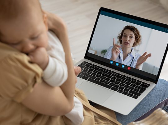 A mother holding a baby talks to a physician on a laptop
